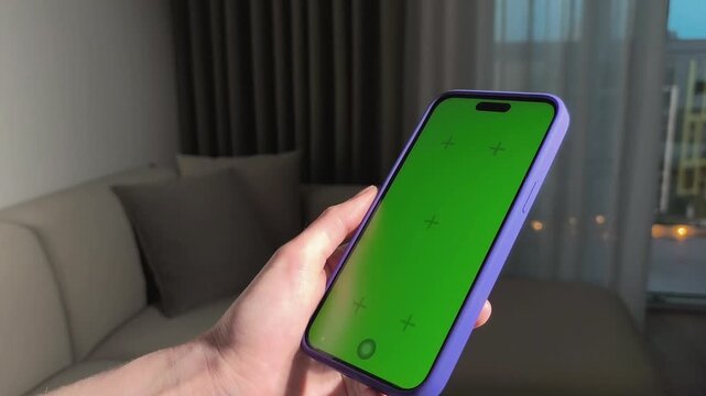 Hands rotate a smartphone with green screen in a living room. The phone shifts from portrait to landscape while held close. Mockup concept for app, advertising, and interface design..