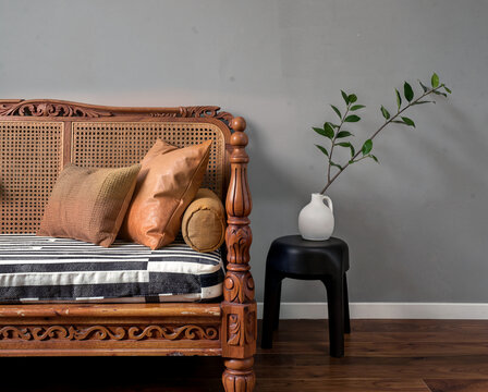 Vintage wooden sofa with cushions beside minimalist black stool holding white vase with green branch against gray wall.