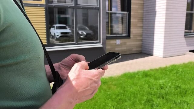 Man taps smartphone outdoors near apartment building. He scrolls on a mobile screen beside green lawn. Casual browsing for modern communication..