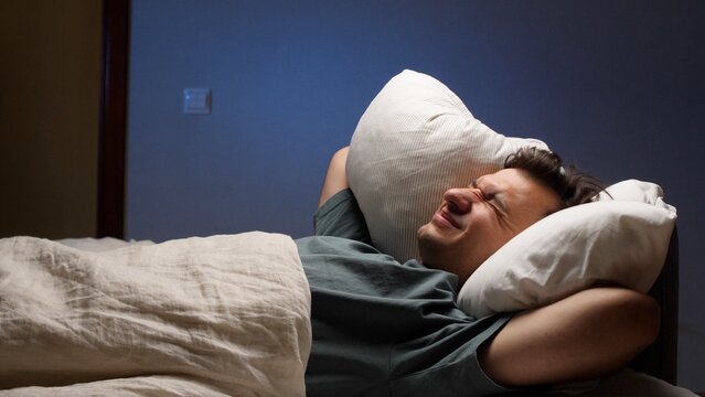 Young man lying in bed experiencing sleep disturbance and insomnia due to noisy neighbors, covering his ears with a pillow to block out the loud sound at night