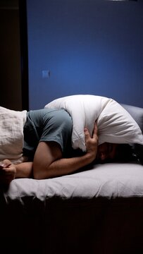 Man finding no sleep in bed, covering his head and ears with a pillow to block out noise, experiencing stress, headache, and fatigue due to a restless night