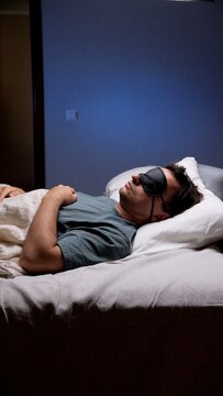 Man sleeping peacefully in a dark bedroom wearing an eye mask, resting comfortably under a duvet for deep, quiet night sleep and improved well being and relaxation
