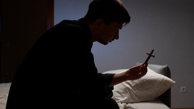 Man in a dim room holding a small wooden cross, quietly praying and reflecting on faith, seeking solace, guidance and spiritual connection in a moment of devotion