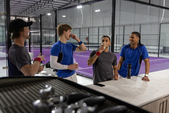 Male teammates in sportswear chatting and drinking from paper cups at racket counter with espresso