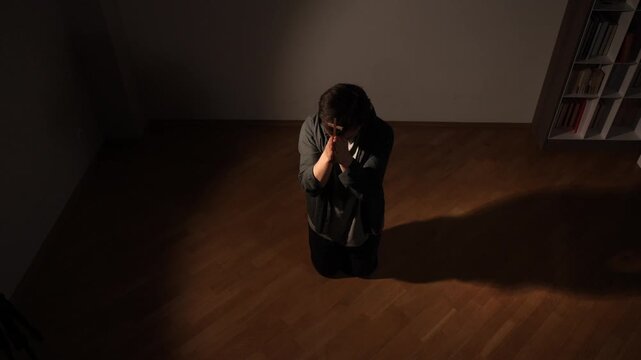 Person kneeling on warm wooden floor in dim room, clasping hands around a cross in quiet prayer, expressing deep devotion, solitude, faith and reverent contemplation