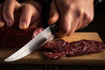 A person uses a knife to slice cured meat on a wooden cutting board in a kitchen. The focus is on the hands and the meat. The scene takes place in the evening with dim lighting © Віталій Б.