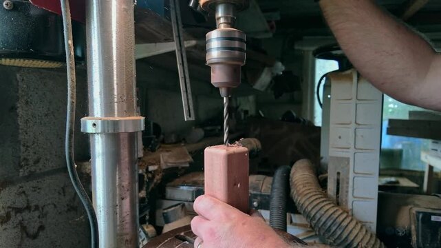 Detailed slow motion view of a craftsman operating a vertical drill press to make a precise hole in a small piece of wood inside a workshop