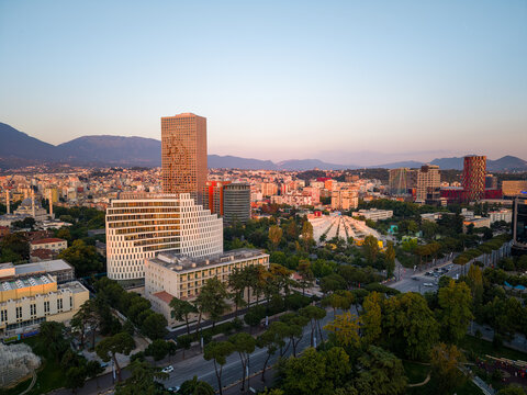 Tirane, Albania - 30 May 2026: Aerial view of the modern skyline featuring skyscrapers, the Pyramid of Tirana, and lush green parks against a backdrop of distant mountains during sunset.