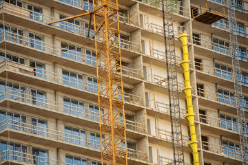 Fototapeta premium Facade of a multi story residential building under construction with installed windows, tower crane and vertical yellow debris chute. Urban development and engineering process.