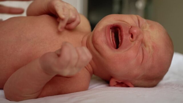 Close up of a distraught newborn baby boy lying down, crying and screaming in agony from abdominal pain and colic, his face red and contorted with discomfort before slowly calming down
