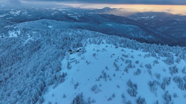 Distant aerial orbit of isolated mountain hut on snowy peak at winter sunset, golden hour wilderness