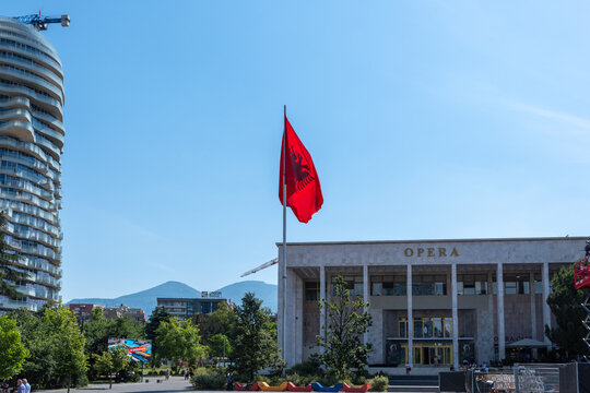 View of the National Theatre of Opera and Ballet with the Albanian flag waving in front of a modern skyscraper under a clear blue sky in Tirana, Tirana County, Albania.