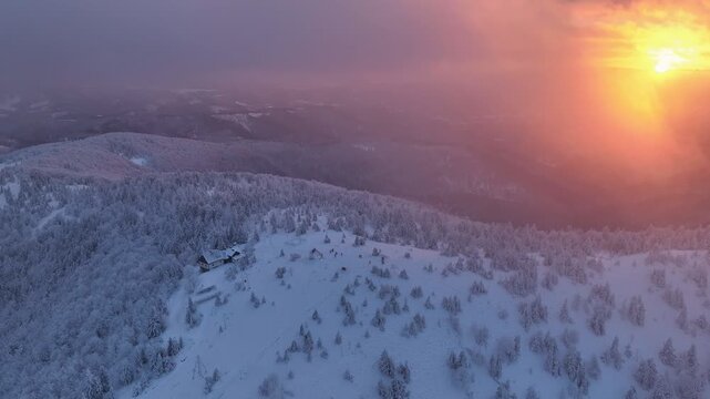 Aerial orbit of snowy mountain summit and hut with sun peaking through diffused sunset clouds