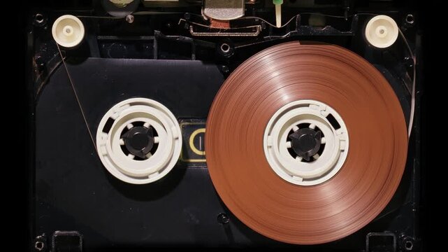Internal mechanism of open retro audio cassette tape playing forward showing brown magnetic tape reels inside black plastic shell against black background