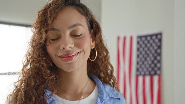 Young woman smiling with hoop earring and blue shirt, american flag visible in a voting station; civic pride.