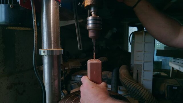 Close up of a persons hands operating a stationary drill press to carefully bore a precise hole longitudinally into a small rectangular piece of wood stock in a workshop environment, Cornwall