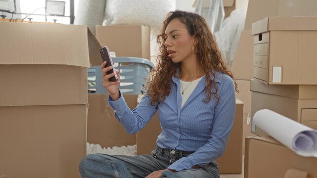 Woman holding smartphone and smiling while seated among stacked moving boxes and packing supplies in a building doorway; new home joy.