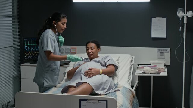 female nurse using stethoscope to check belly of pregnant Black woman in hospital bed. Video is ideal for maternity care and prenatal healthcare marketing