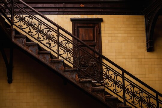 Rustic iron staircase with ornate railing leads to dark wooden door. Yellow tiled wall and dark wooden trim create a vintage interior atmosphere. Abandoned industrial building.