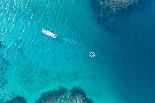 Aerial view of a small white boat towing a round inflatable tube with people across clear turquoise waters and dark coral reefs Naviti, Western Division, Fiji.