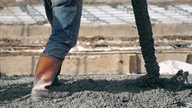 Close up of a construction worker in muddy rubber boots wading through fresh wet concrete near wooden formwork panels during a floor casting process