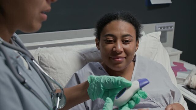 Black pregnant woman lying in hospital bed while nurse measuring her temperature with infrared thermometer. Clip is ideal for maternity care and prenatal health, over shoulder shot