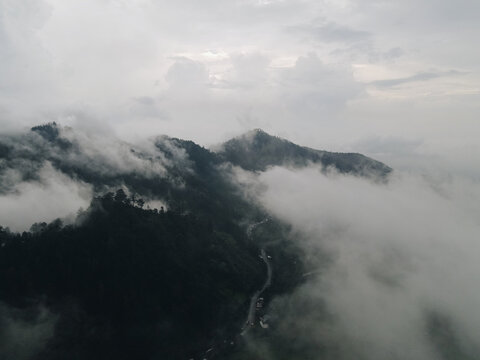 Aerial view of misty mountains and lush green slopes partially covered by thick white clouds with a winding road below under an overcast sky in Tawangmangu, Central Java, Indonesia.