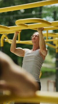 A muscular man is intensely performing pull-ups on yellow monkey bars at an outdoor fitness park. He demonstrates strength and dedication during his workout routine