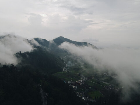 Aerial view of misty mountains and a valley village partially covered by thick white clouds under an overcast sky in Tawangmangu, Central Java, Indonesia.