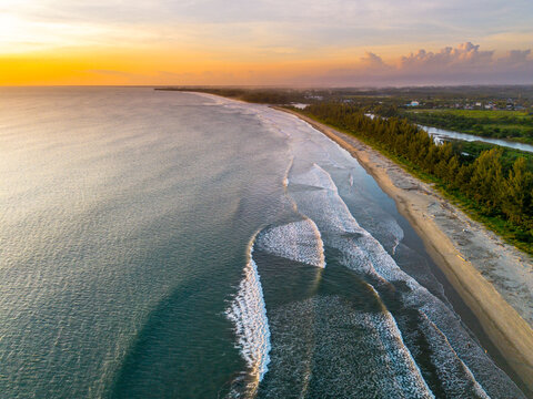 Aerial view of the long sandy coastline with rolling waves, lush green trees, and a golden sunset sky over the ocean in Bengkulu, Bengkulu, Indonesia.
