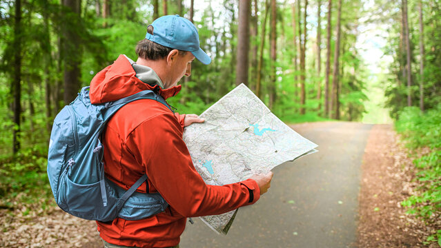 Hiker looking at a map on a forest path, wearing a orange waterproof jacket and backpack, holding a paper map with trails.