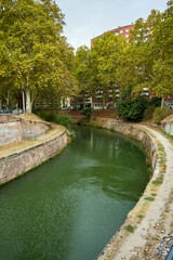 Fototapeta premium A narrow, tree-lined canal curves through a quiet urban area with stone embankments and a dirt path. Buildings rise behind dense green foliage; canal.