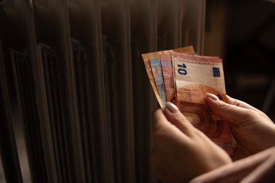 Woman holding euro notes next to a radiator, illustrating rising heating costs, household budget strain and energy crisis impact on personal finances and winter comfort