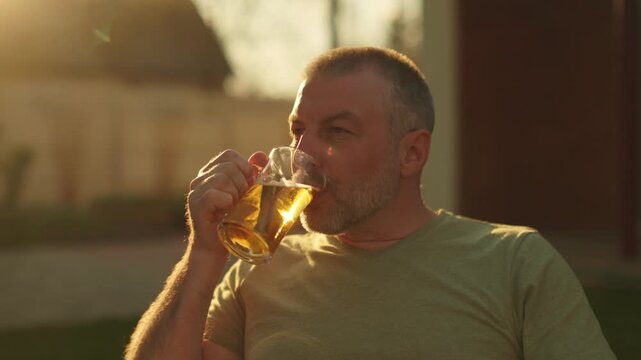 Happy middle-aged man drinking lager beer from mug and smiling in backyard, 4k