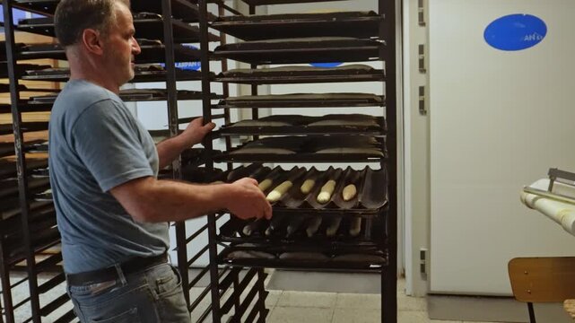 Professional baker arranging uncooked baguette dough on a tray. He places the tray on a large metal rack to let the bread rise before baking
