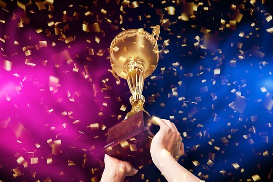 Gold trophy cup held by hands with falling confetti and colorful lights during a victory celebration