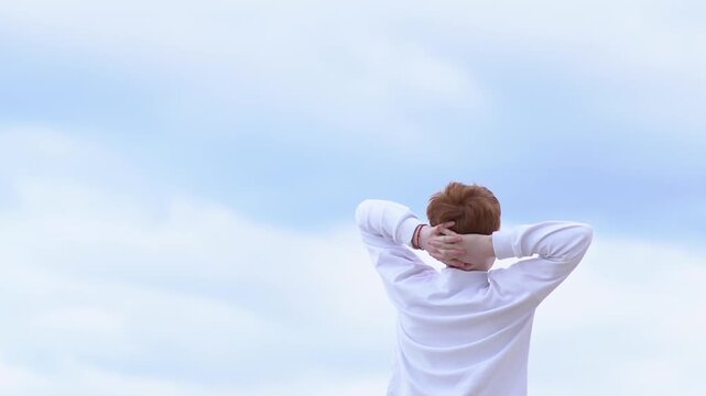 A relaxed teenage boy breathes in the fresh air with his hands behind his head, basking in the sun. Dreams, freedom, and the concept of travel. High quality FullHD footage