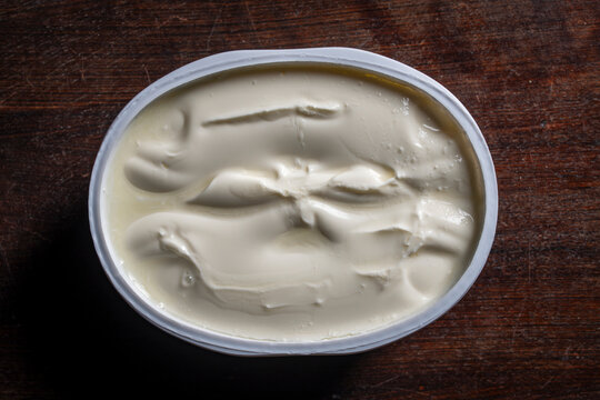 Creamy white sour cream or processed cheese in plastic container on wooden table, closeup. Top view of thick white yogurt cream cheese or sour cream inside an oval plastic cup on dark background