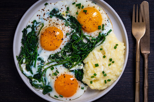Fried eggs with greens and mashed potatoes for breakfast, close up. Top view of healthy plate of three fried eggs with green spinach and creamy mashed potatoes sprinkled with chives