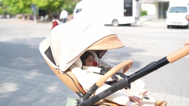 Soft beige stroller carries sleeping child. Sunlit street with distant vehicles and green trees. Child rests peacefully, wrapped in cozy blanket. Ideal for parenting, travel, or lifestyle imagery