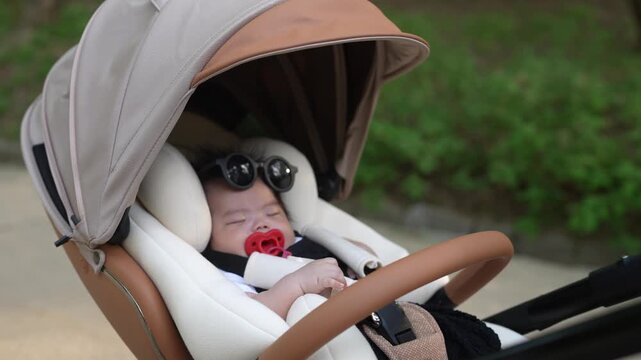 Baby sleeps peacefully in stroller. Red pacifier rests in baby's mouth. Baby wears sunglasses, hands on steering wheel. Stroller has beige canopy and wooden handle