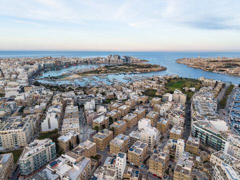 Aerial view of dense urban residential buildings and a harbor filled with yachts under a clear blue sky in Malta.