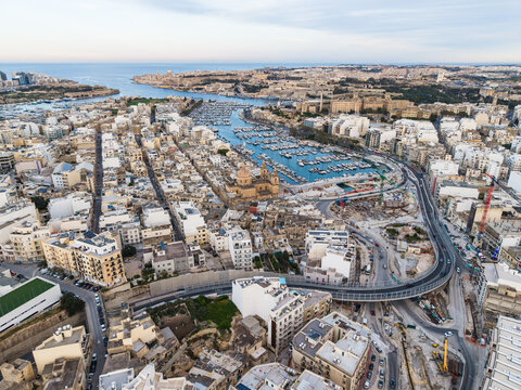 Aerial view of the Msida marina and Saint Joseph Parish Church with dense urban buildings and a modern flyover bridge under a clear sky in Msida, Eastern Region, Malta.