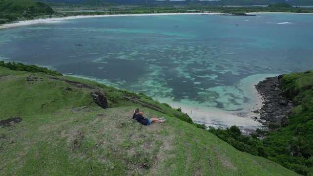 Caucasian Woman Resting On Grassy Cliff Overlooking Turquoise Bay, Panoramic Aerial View Of Coral Reef Patches And Calm Water, Solo Traveler Lying Back, Bukit Merese, Lombok, Tanjung Ann