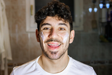 Obraz premium Young man applying facial cream and smiling, morning skincare routine and self-care