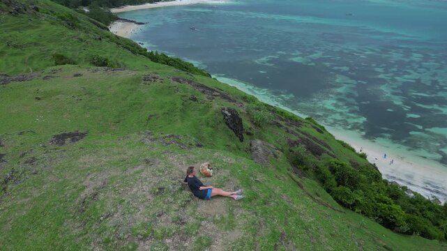 Caucasian Woman Resting On Grassy Cliff Overlooking Turquoise Bay, Panoramic Aerial View Of Coral Reef Patches And Calm Water, Solo Traveler Lying Back, Bukit Merese, Lombok, Tanjung Ann