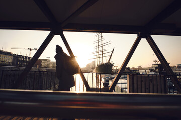 Naklejka premium A lone figure stands on a metal-framed pedestrian bridge overlooking a harbor at sunset. Backlit by the low sun, the person is rendered as a dark silhouette