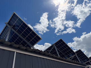 Obraz premium Angled rooftop array of photovoltaic panels mounted on metal frames above a corrugated roof, photographed from below with a vivid blue sky and scattered white