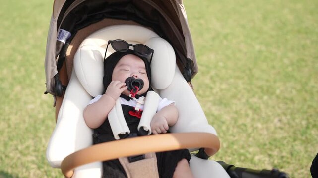 Soft sunlight bathes green grass and stroller. Baby sleeps peacefully, wearing sunglasses. Parent gently adjusts stroller, offering calm care. Ideal for family, parenting, or outdoor lifestyle themes