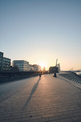 Obraz premium A wide paved promenade beside a city harbor at sunset, with low sunlight casting long silhouettes of a lone cyclist and distant pedestrians. Modern waterfront